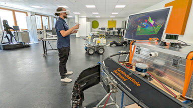 Eine Person mit einer VR-Brille steht in einem Labor. Vor ihr ein Monitor mit bunten Linien.