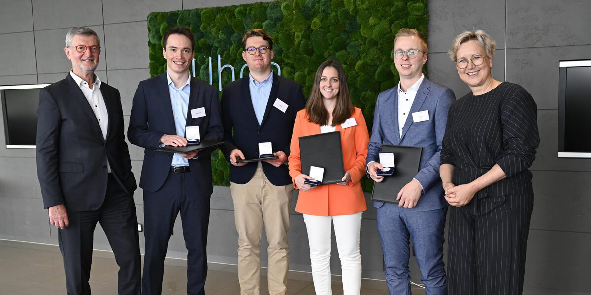 Sechs Personen stehen in einer Reihe nebeneinander, die mittleren vier halten eine Mappe und ein Schachtel mit einer Medaille vor sich.