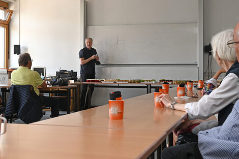 Eine männlich gelesene Person steht in einem Seminarraum an einer Tafel und erklärt. An den Tischen sitzen ältere Menschen.