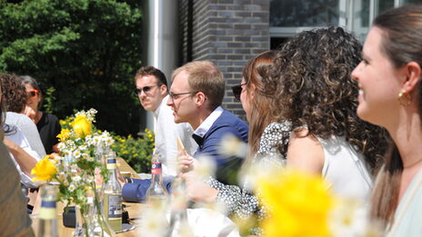 View of the long table decorated with flowers set up in front of the building, where former students are chatting animatedly.