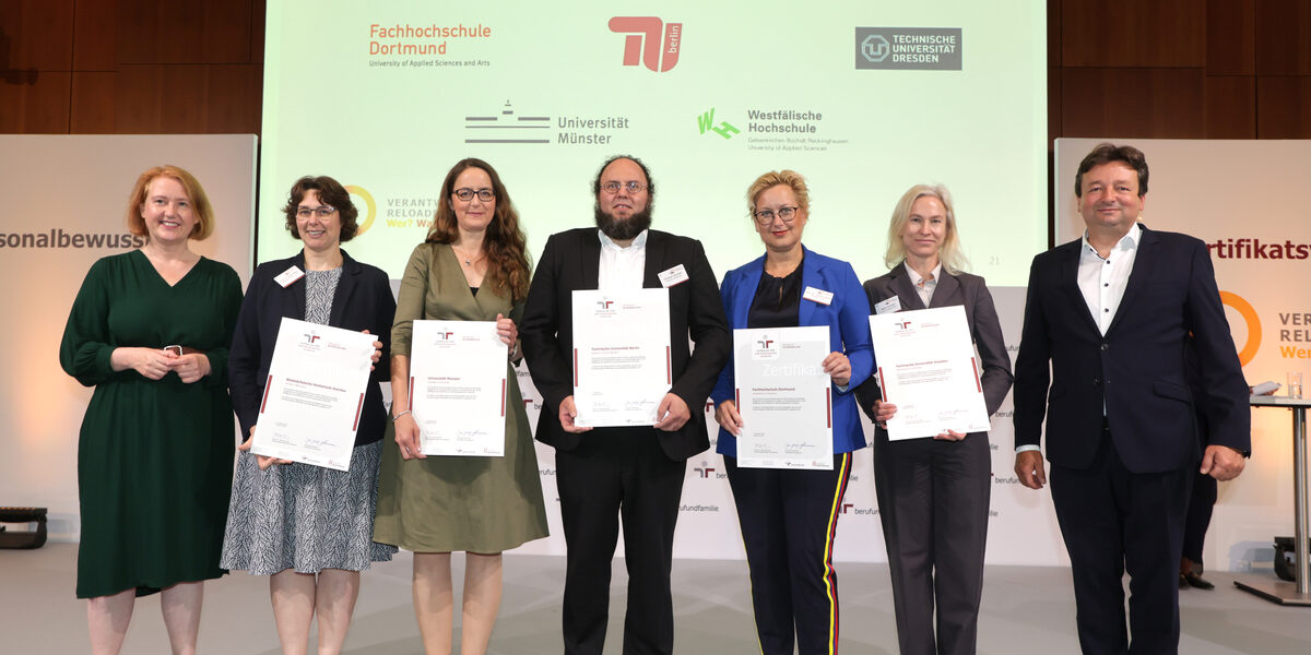 Seven people stand next to each other on a stage, five holding a certificate in front of them. The logos of Fachhochschule Dortmund, TU Berlin, TU Dresden, Münster University and Westfälische Hochschule can be seen on a screen in the background.