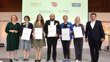 Sieben Personen stehen nebeneinander auf einer Bühne, fünf halten ein Zertifikat vor sich. Im Hintergrund auf einer Leinwand zu sehen sind die Logos der Fachhochschule Dortmund, der TU Berlin, der TU Dresden, der Uni Münster und der Westfälischen Hochschule.