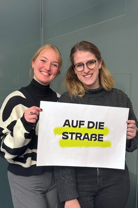 Zwei Personen stehen nebeneinander und halten ein Schild mit der Aufschrift "Auf die Straße".