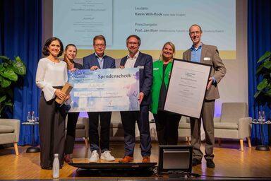 Six people stand next to each other on a stage. They present a large-format donation check and a certificate.