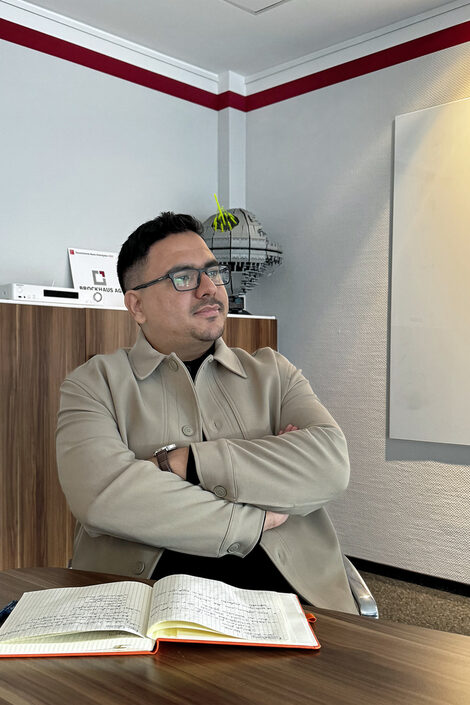 Eine Person sitze an einem Konferenztisch und hört zu. Vor ihr liegt ein aufgeschlagenes Notizbuch.