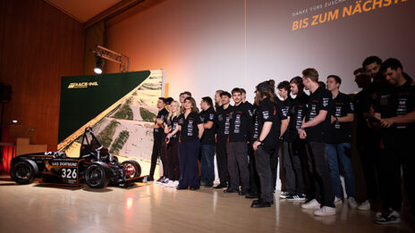 Around 20 people in black T-shirts are standing on a stage, with a racing car to their left.