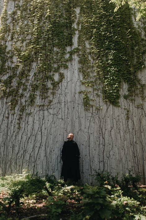 A man in black clothes stands with his back against an ivy-covered rock face. His face is turned upwards to the right. The ground in the foreground is overgrown with low bushes.