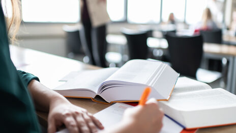 Photo of a woman taking notes. There are two books in front of her. In the background are several people in the seminar room. __ Woman takes notes. There are two books in front of her. In the background there are several people in the seminar room.