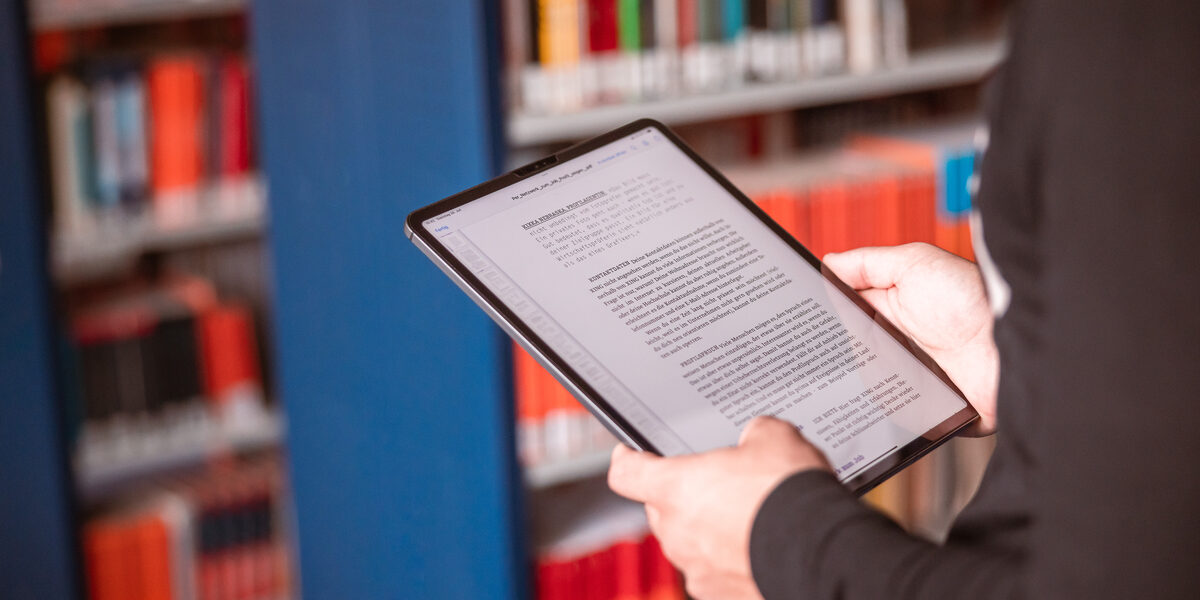 Photo of a tablet in the hand of someone standing in a library.