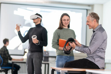 Foto von einem Lehrenden und einer Studierenden. Der Lehrende zeigt der Studierenden eine VR-Brille. Zentral sieht man außerdem einen Studierenden, der eine VR-Brille trägt und diese bedient. Im Hintergrund sitzt ein Studierender an einem PC vor einer großen Leinwand.