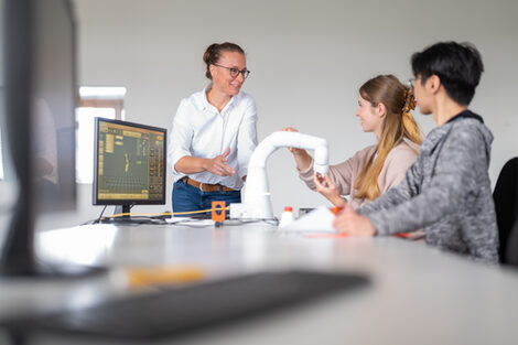 Eine Professorin steht an einem Tisch, an dem ein Roboterarm aufgebaut ist und ein Student und eine Studentin arbeiten. Sie erklärt den beiden etwas dazu. __A professor stands at a table where a robot arm is set up and a male and female student are working. She explains something about it to the two of them.<br><br>