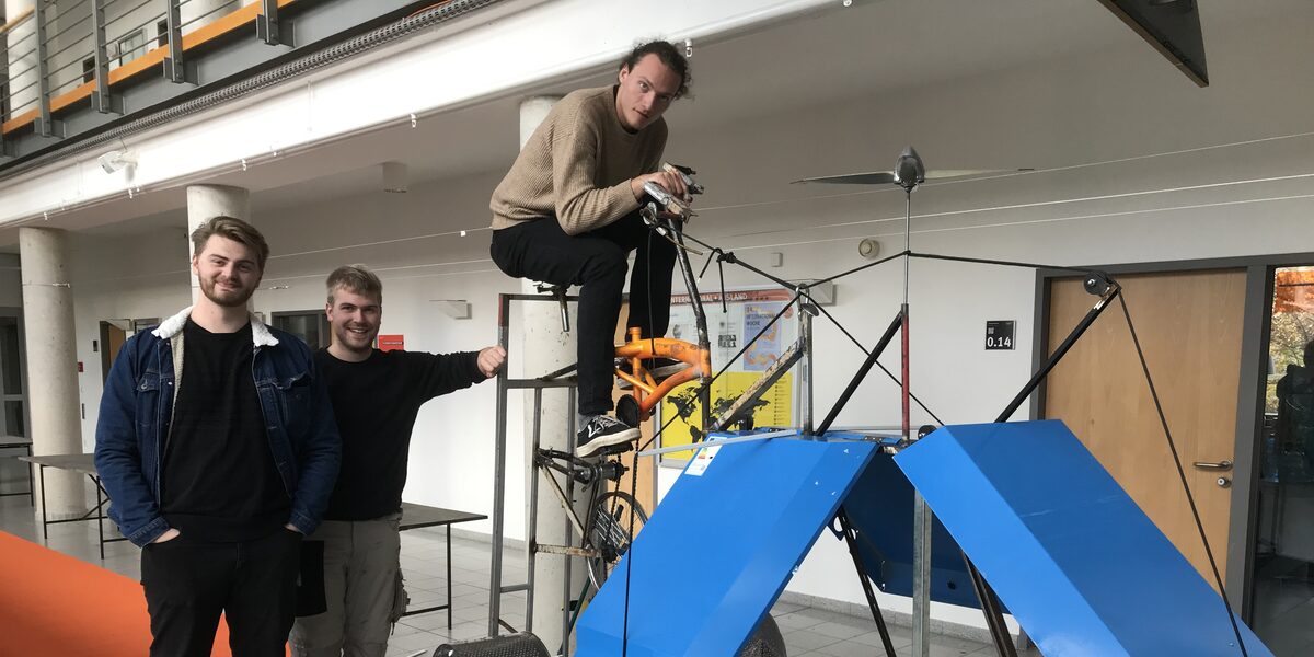 Three students stand or sit on a self-built machine.