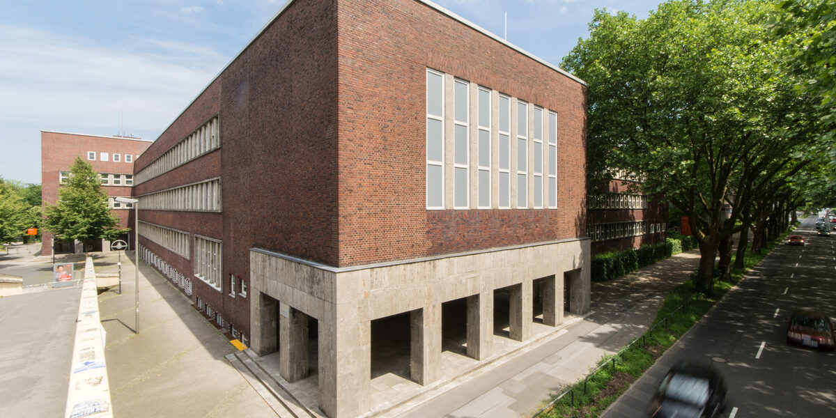 Photo with a view of parts of a building of the Fachhochschule Dortmund at Max-Ophüls-Platz with forecourt and sidewalk as well as street with cars next to it.