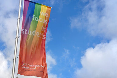 Eine FH-Dortmund-Fahne mit Regenbogen weht vor blauem Himmel.