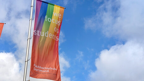Eine FH-Dortmund-Fahne mit Regenbogen weht vor blauem Himmel.