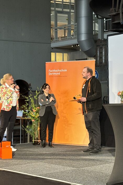 Three people are standing on a stage. One person is holding an award.