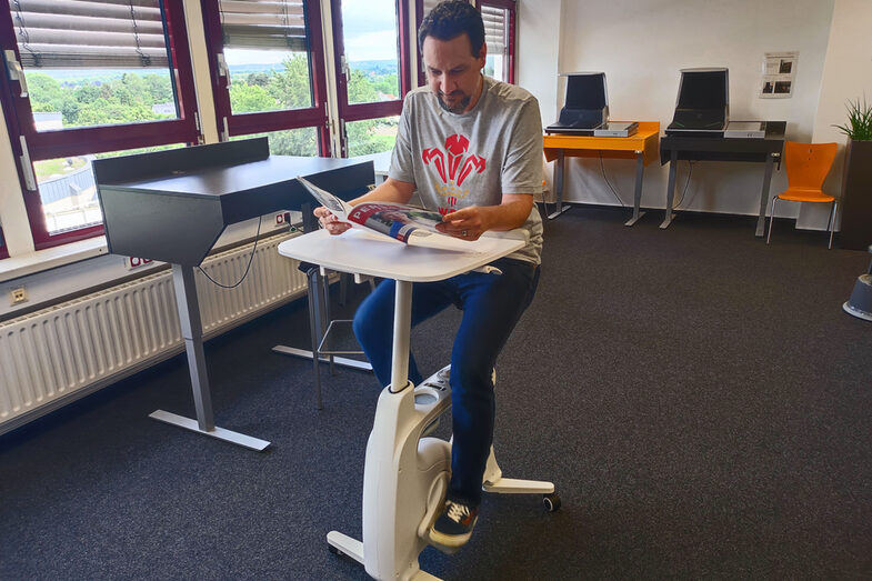 In der Mitte eines Raumes steht ein fahrradähnliches Gerät, allerdings ohne Räder und ohne Lenker, dafür mit einem kleinen Tisch. Auf dem Gerät sitzt eine Person, tritt in die Pedale und liest in einer Zeitschrift.