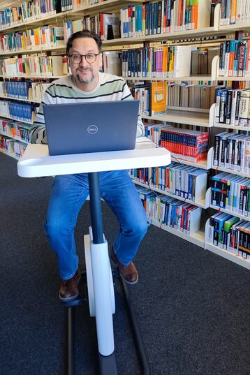 Foto von einem Mann auf einem Indoor-Fahrrad mit kleinem Tisch, auf dem ein Laptop steht__Photo of a man sitting on an indoor bicycle with a small table, a notebook is placed on the table