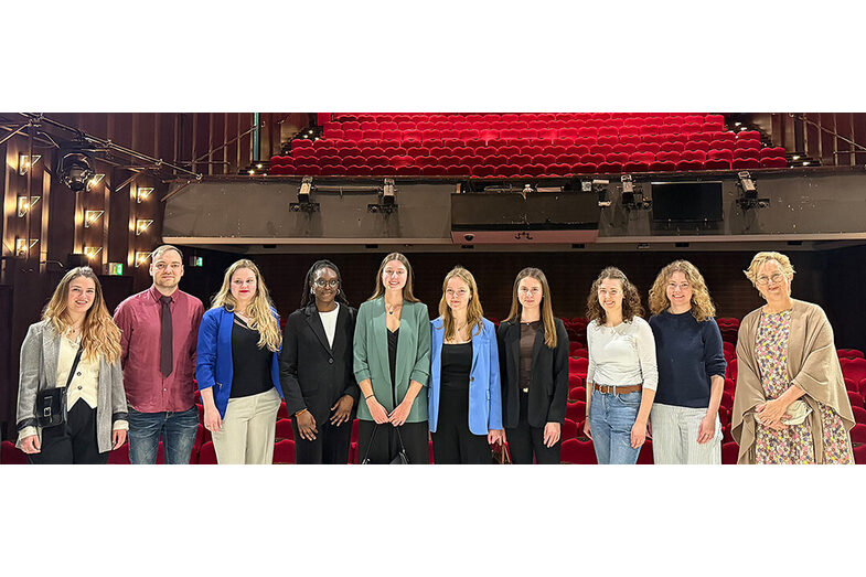 Mehrere Personen stehen nebeneinander in einem Saal, hinter ihnen sind rot gepolsterte leere Sitzreihen zu sehen.