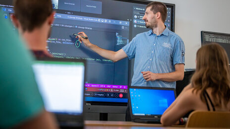 Photo of a lecture situation in a seminar room. A man is standing in front of a digital board and explaining something in code.