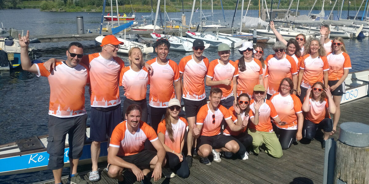 20 people in identical jerseys stand or squat on a jetty in front of a long canoe floating on the lake behind them.