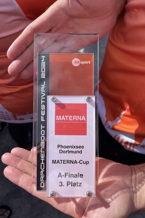 Zwei Hände halten einen Pokal aus Plexiglas mit der Aufschrift "Drachenboot Festival 2024, Phoenixsee Dortmund, Materna-Cup, A-Finale, 3. Platz".