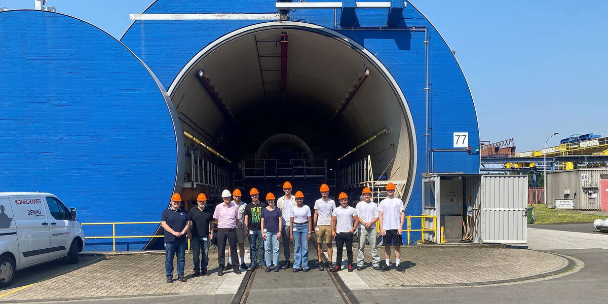 Twelve people, each wearing a hard hat, stand in front of the entrance to a large round hall.