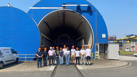 Twelve people, each wearing a hard hat, stand in front of the entrance to a large round hall.