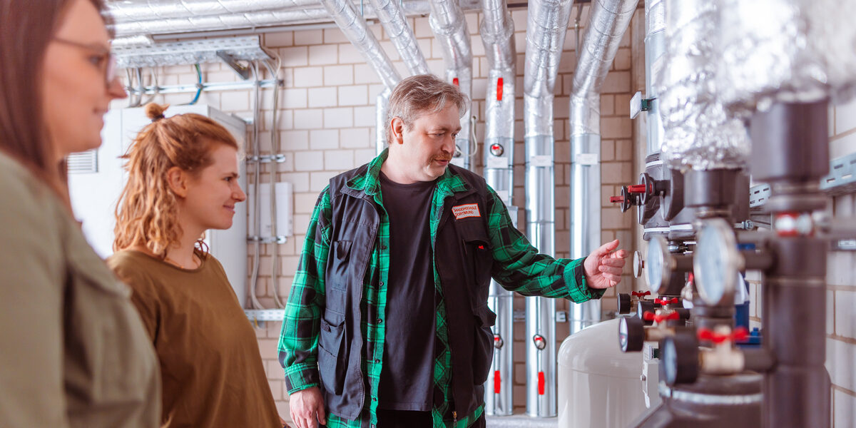 Photo of 3 people standing in front of a heating system. The man seems to be explaining something to the two women.