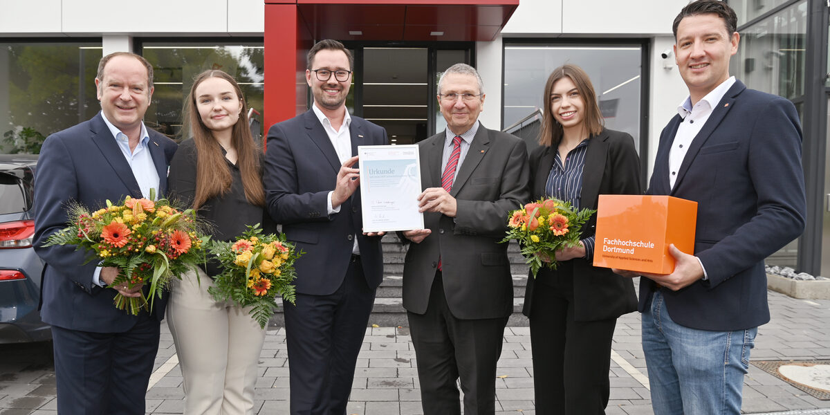 Sechs Personen stehen nebeneinander, blicken freundlich in dei Kamera, im Hintergrund eine kurze Treppe zum Eingang eines Geschäfts mit dem Wort "Weckbacher" auf dem Vordach. Drei Personen halten einen Blumenstrauß in den Händen, die beiden mittleren halten zusammen eine Urkunde in die Kamera.