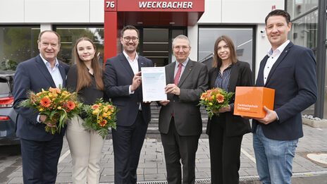 Sechs Personen stehen nebeneinander, blicken freundlich in dei Kamera, im Hintergrund eine kurze Treppe zum Eingang eines Geschäfts mit dem Wort "Weckbacher" auf dem Vordach. Drei Personen halten einen Blumenstrauß in den Händen, die beiden mittleren halten zusammen eine Urkunde in die Kamera.