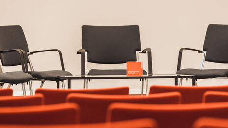 Foto eines leeren Veranstaltungsraums, Blick über leere Stuhlreihen hinweg zum Podium, wo weitere Stühle stehen sowie ein Tisch mit FH-Würfel. __ Empty event room, view over empty rows of chairs to the podium, where there are more chairs and a table with FH cube.