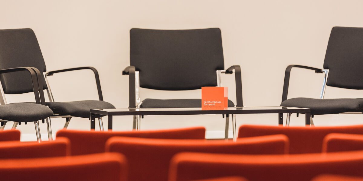 Foto eines leeren Veranstaltungsraums, Blick über leere Stuhlreihen hinweg zum Podium, wo weitere Stühle stehen sowie ein Tisch mit FH-Würfel. __ Empty event room, view over empty rows of chairs to the podium, where there are more chairs and a table with FH cube.