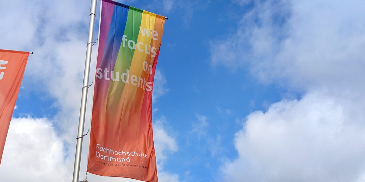 Eine FH-Dortmund-Fahne mit Regenbogen weht vor blauem Himmel.