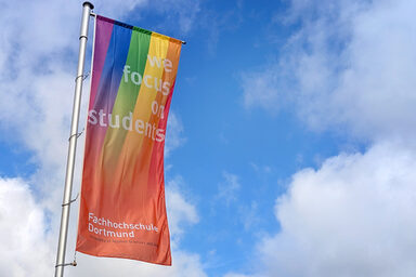 Eine FH-Dortmund-Fahne mit Regenbogen weht vor blauem Himmel