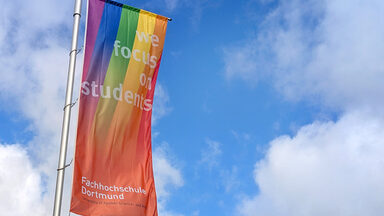 Eine FH-Dortmund-Fahne mit Regenbogen weht vor blauem Himmel