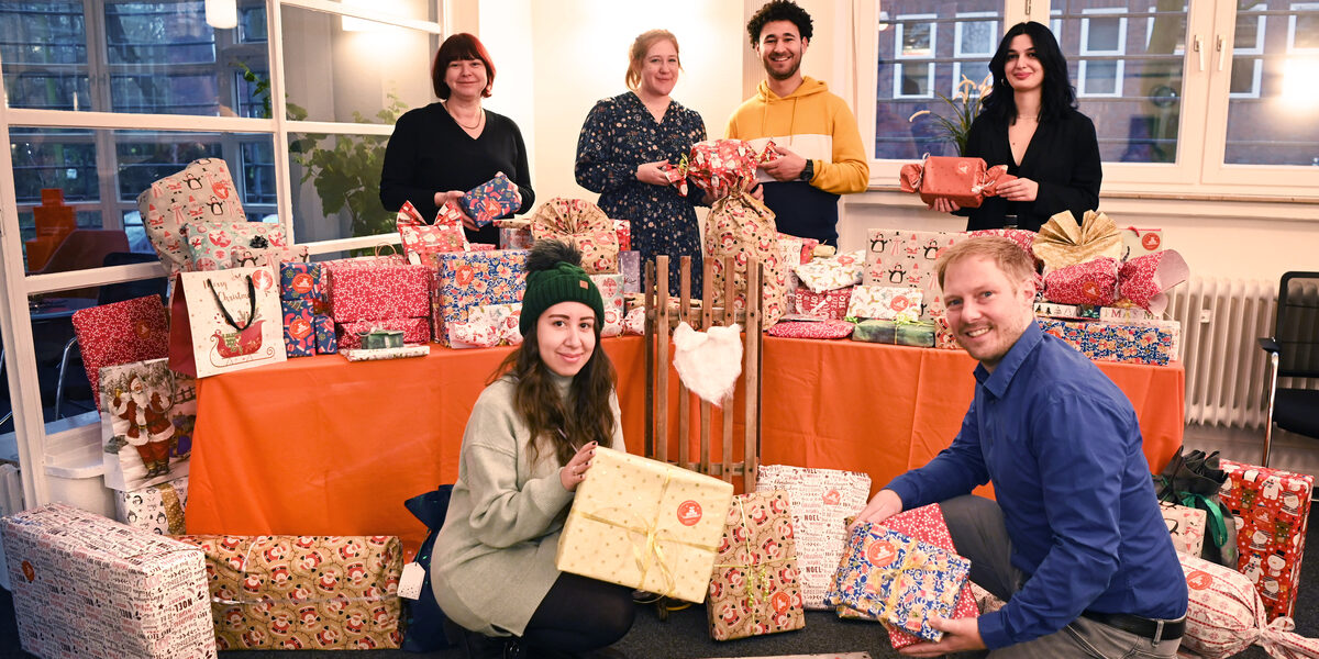 Hinter einem orange gedeckten Tisch mit vielen eingepackten Geschenken stehen vier Personen, die ihrerseits Geschenke in den Händen halten. Vor dem Tisch hocken zwei weitere Personen. Alle blicken lächelnd zur Kamera.