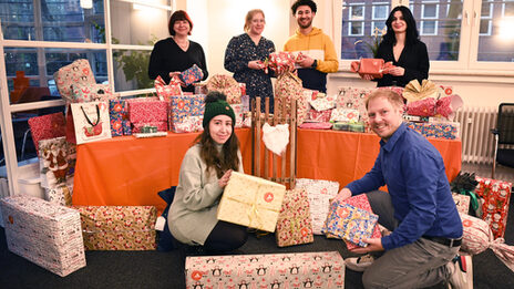 Hinter einem orange gedeckten Tisch mit vielen eingepackten Geschenken stehen vier Personen, die ihrerseits Geschenke in den Händen halten. Vor dem Tisch hocken zwei weitere Personen. Alle blicken lächelnd zur Kamera.