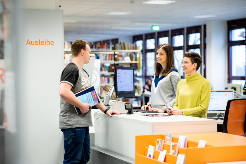 Foto von zwei Mitarbeiterinnen, die hinter der Theke der Bibliothek stehen und mit dem ihnen gegenüber stehenden Studierenden, der Bücher in der Hand hat, im Gespräch sind.