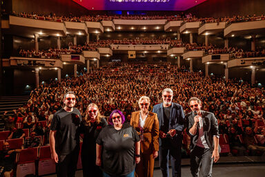 Several people wearing sunglasses on stage in the opera house. In the background, the first-semester students in the stands make "la Ola".