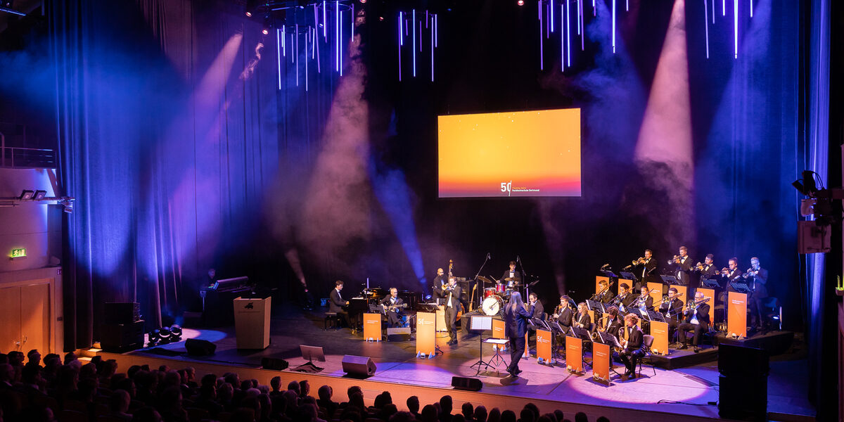 Auf der Bühne im Konzerthaus spielt eine Big-Band. Die Notenständer sind mit "50 Jahre Fachhochschule Dortmund" verhüllt. Die Bühne erstrahlt in orangefarbenem Licht.