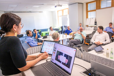 Photo of an event in a seminar room. The focus is on a young lady at the lectern, where there is also a laptop, listening intently to the speaker in the group.