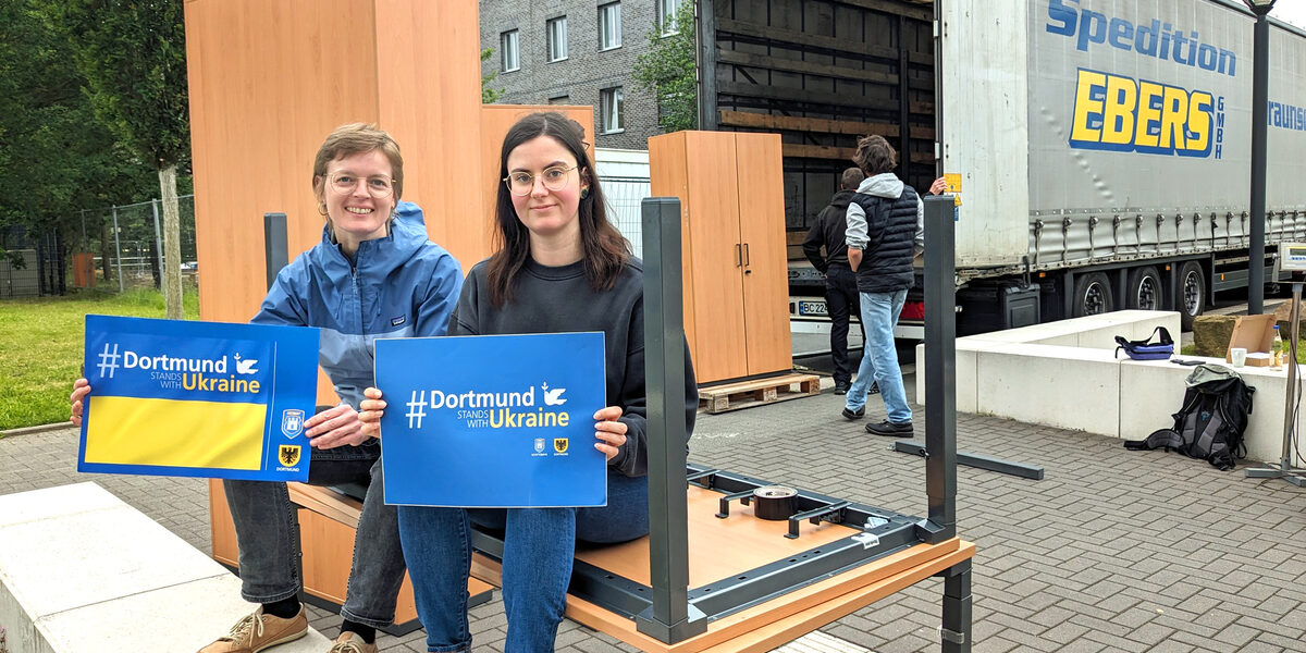 Zwei Personen sitzen auf einem Schreibtisch und halten Schilder mit der Aufschrift "Dortmund stands with Ukraine" in der Hand. Im Hintergrund werden Möbel auf einen Lkw verladen.