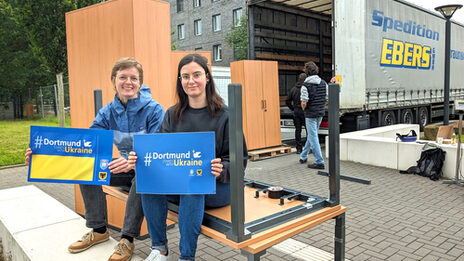 Zwei Personen sitzen auf einem Schreibtisch und halten Schilder mit der Aufschrift "Dortmund stands with Ukraine" in der Hand. Im Hintergrund werden Möbel auf einen Lkw verladen.