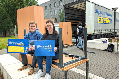 Two people are sitting on a desk holding signs reading "Dortmund stands with Ukraine". In the background, furniture is being loaded onto a truck.