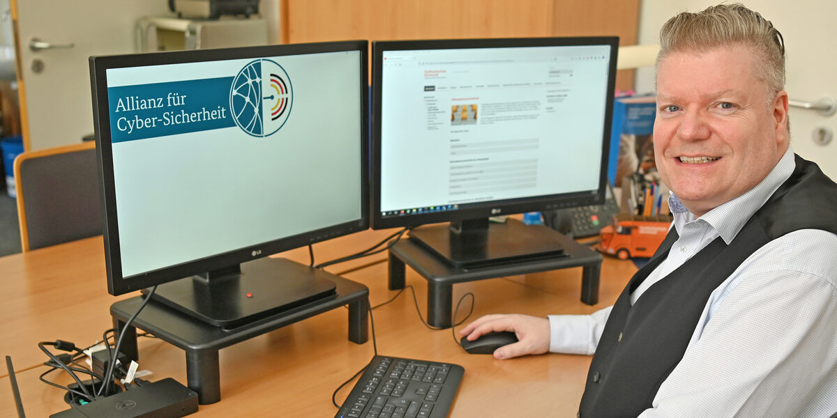 A male person is sitting at a desk in front of two monitors. The logo of the Alliance for Cyber Security can be seen on one monitor.