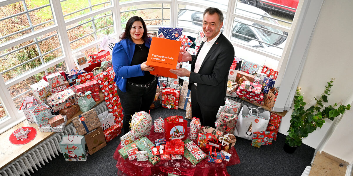 Zwei Personen stehen inmitten von weihnachtlich verpackten Geschenken und halten gemeinsam einen orangefarbenen FH-Dortmund-Würfel in den Händen.