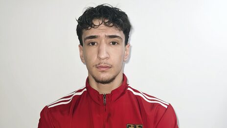 Ein Mensch in roter Trainingsjacke mit dem deutschen Wappen auf der Brust schaut mit verschränkten Armen in die Kamera.