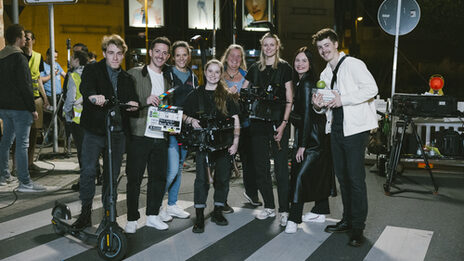 A group of people are standing on a crosswalk, one person is holding an e-scooter, two other people are holding a film camera and a film clapperboard.