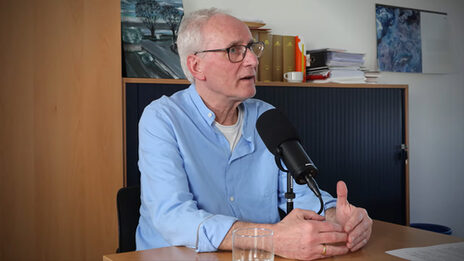 Eine Person sitzt an einem Tisch vor einem Mikrofon und spricht.